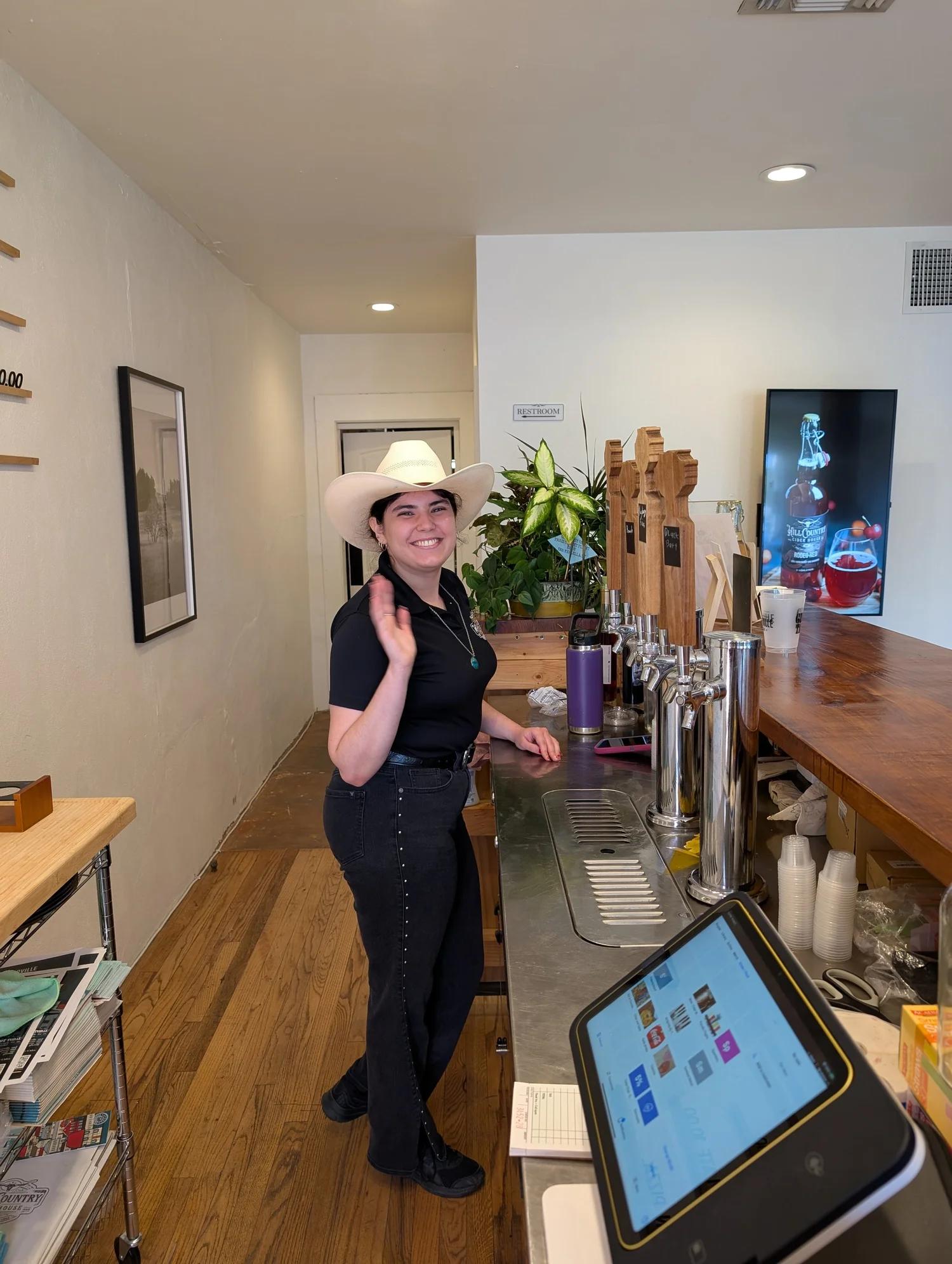 A smiling host in a white cowboy hat behind the bar at Hill Country Cider House